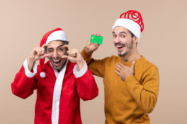 Vista frontal de dois caras felizes, um com casaco de Papai Noel e o outro com crédito, colocando a mão no peito em um fundo bege isolado