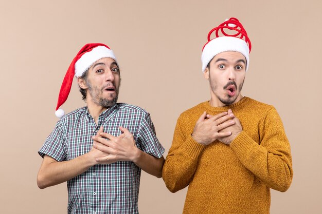 Vista frontal de dois caras surpresos com chapéus de Papai Noel colocando as mãos no peito em um fundo bege isolado