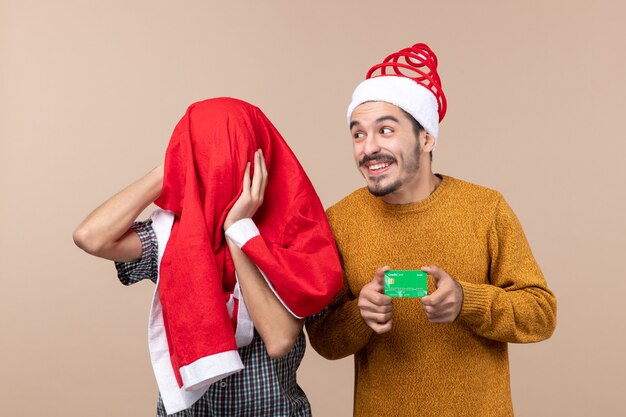 Vista frontal de dois caras, um segurando um cartão de crédito e o outro cobrindo a cabeça com um casaco de Papai Noel em fundo bege isolado