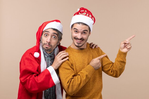 Vista frontal de dois homens de natal com chapéu de Papai Noel, um segurando o ombro do amigo e o outro mostrando a direção em um fundo bege isolado