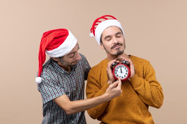 Vista frontal de dois homens de natal, um segurando um despertador com os olhos fechados e o outro mostrando as horas em um fundo bege isolado