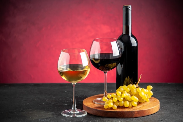 Vista frontal de duas taças de vinho uvas amarelas em uma garrafa de vinho de placa de madeira no fundo vermelho