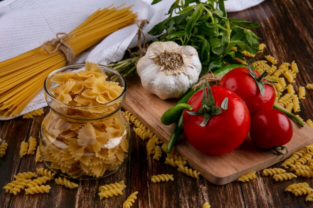 Vista frontal de espaguete cru com macarrão em uma jarra com tomate, alho e pimenta em uma placa de corte e com um punhado de hortelã em uma superfície de madeira