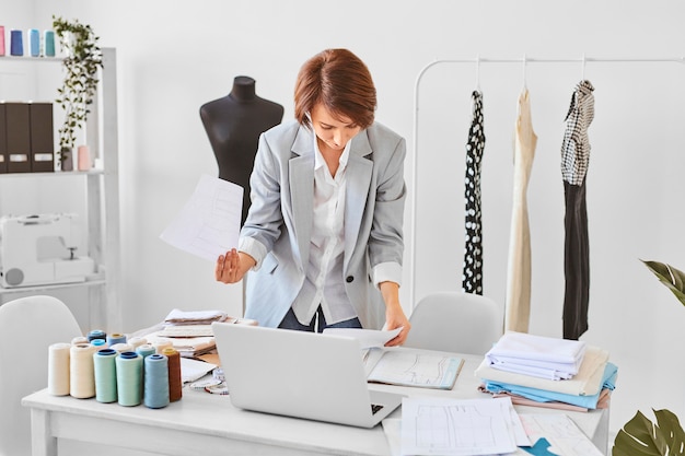 Vista frontal de estilista feminina, consultando planos de linha de roupas no ateliê