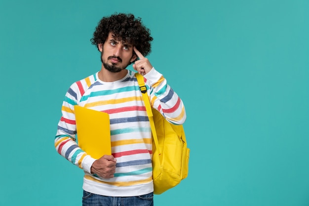 Vista frontal de estudante do sexo masculino com camisa listrada e mochila amarela segurando arquivos pensando na parede azul