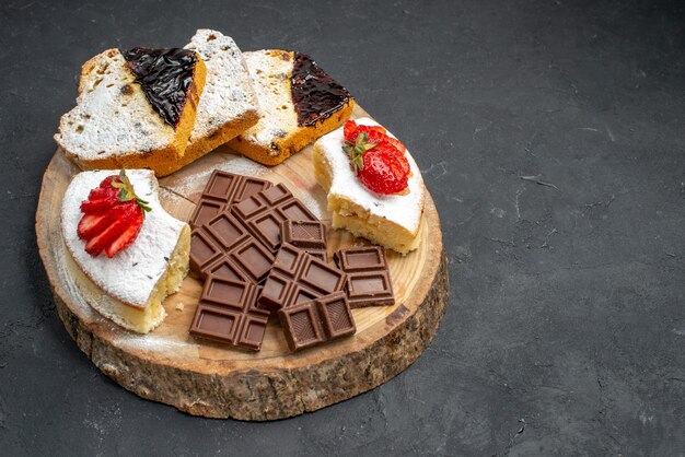 Vista frontal de fatias de bolo saboroso com frutas e barras de chocolate em fundo escuro