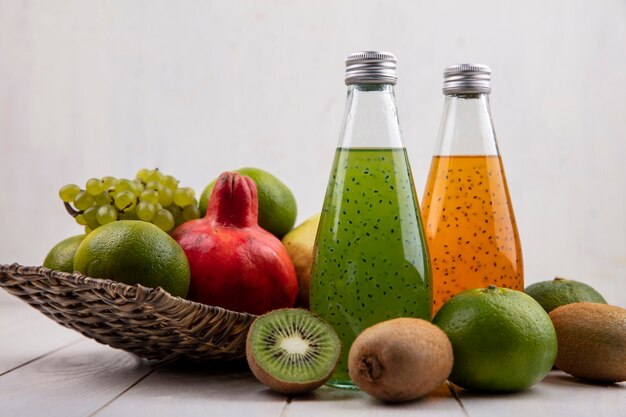 Vista frontal de garrafas de suco com romãs, uvas, tangerinas e peras em uma cesta em uma parede branca