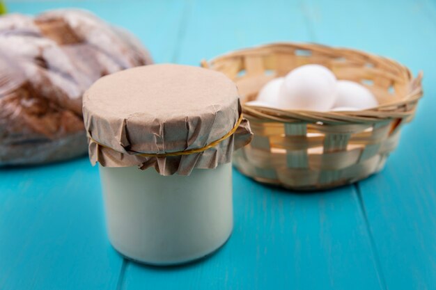 Vista frontal de iogurte em uma jarra com ovos de galinha em uma cesta e pão preto em um fundo turquesa