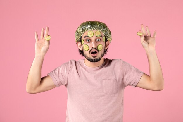 vista frontal de jovem aplicando máscara de pepino no rosto na parede rosa
