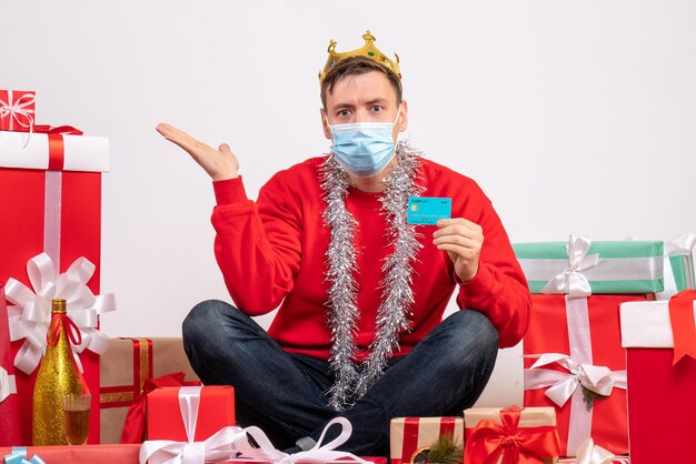 Vista frontal de jovem com máscara sentado em volta de presentes de Natal com cartão do banco na parede branca