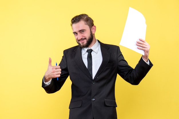 Vista frontal de jovem de terno preto segurando papel em branco sobre amarelo com sorriso, polegar para cima com dedos, excelente sinal