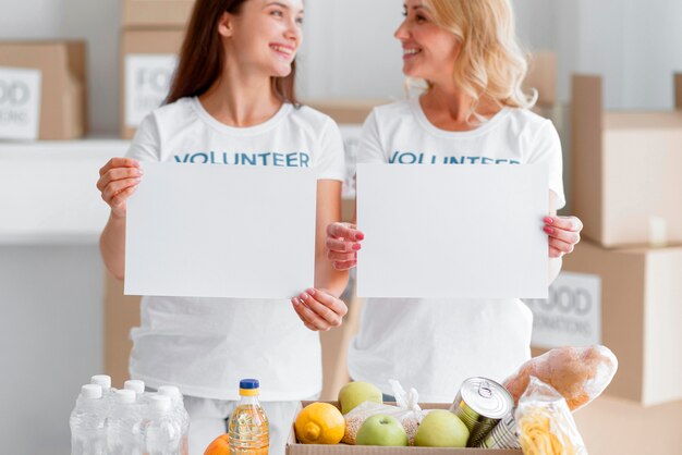 Vista frontal de mulheres voluntárias sorridentes posando com cartazes em branco e doações de alimentos