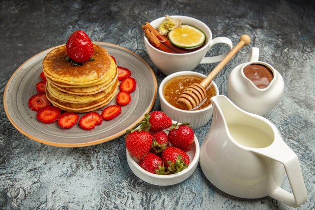Vista frontal de panquecas deliciosas com frutas e uma xícara de chá na superfície clara de bolo doce de frutas