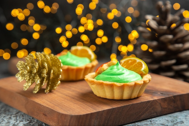 Vista frontal de pequenas tortas na tábua de cortar enfeites de natal em uma superfície escura com luzes de natal