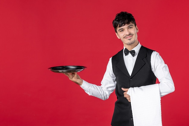 Vista frontal de perto de jovem garçom sorridente confiante e feliz em um uniforme com uma borboleta no pescoço segurando a bandeja e uma toalha em fundo vermelho