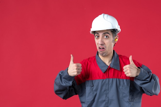 Foto grátis vista frontal de perto de jovem trabalhador confiante em uniforme, com capacete e tampões de ouvido, fazendo gesto de ok na parede vermelha isolada