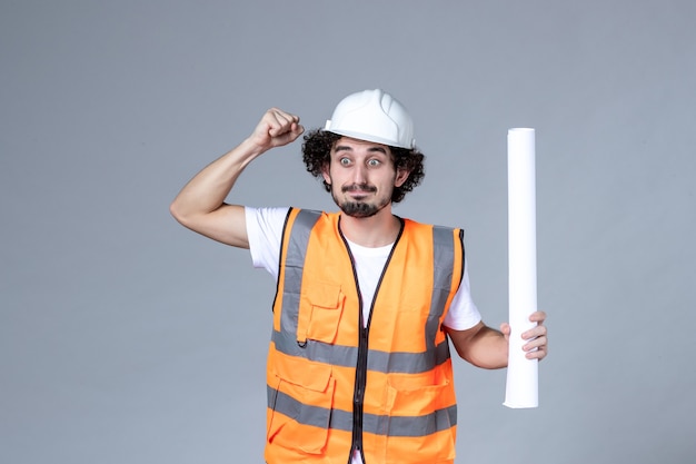 Vista frontal de perto de nervoso emocional trabalhador da construção civil masculino em colete de advertência com capacete de segurança e segurando em branco na parede cinza