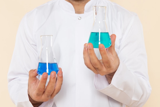 Vista frontal de perto jovem químico masculino em terno especial branco segurando frascos com soluções na parede creme experimento científico laboratório de química