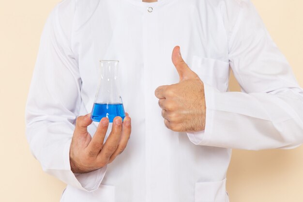 Vista frontal de perto jovem químico masculino em terno especial branco segurando o pequeno frasco com solução azul na parede creme experimento científico.