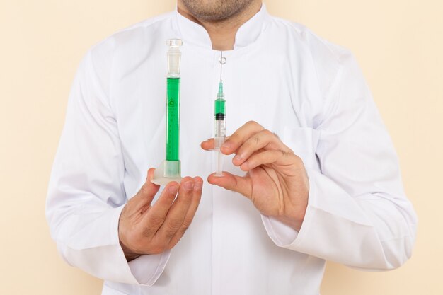 Vista frontal de perto jovem químico masculino em terno especial branco segurando o pequeno frasco com solução verde e injeção no laboratório de experimentos científicos de parede creme