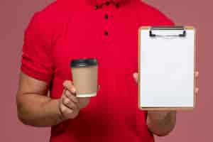 Foto grátis vista frontal de perto mensageiro de uniforme vermelho segurando a xícara de café de entrega e o bloco de notas na parede rosa trabalho masculino trabalhador