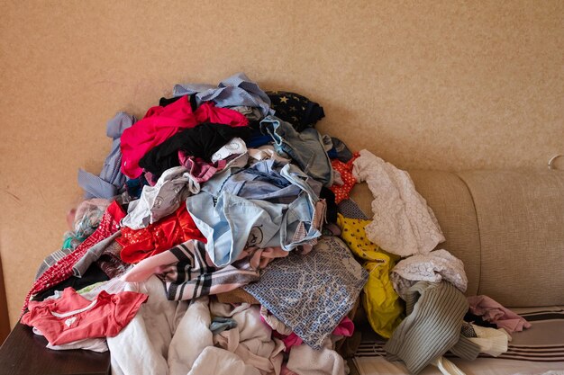 Vista frontal de roupas infantis coloridas espalhadas no sofá em casa Grande monte de roupas deitadas no quarto antes da lavanderia Conceito de limpeza de lama de bagunça e tarefas domésticas