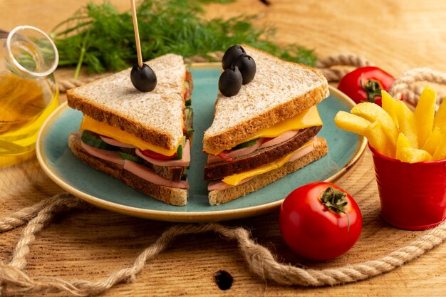 Vista frontal de sanduíches saborosos com tomate verde-oliva e presunto no prato junto com batatas fritas e tomate com óleo na madeira