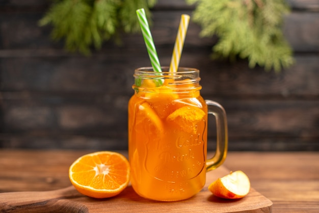 Vista frontal de suco de frutas frescas em um copo servido com tubos, maçã e laranja em uma tábua de madeira sobre uma mesa marrom