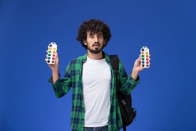 Foto grátis vista frontal de um aluno do sexo masculino com camisa xadrez verde e mochila preta segurando tintas para desenhar na parede azul