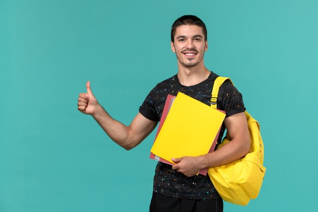 Vista frontal de um aluno do sexo masculino em uma mochila de camiseta amarela escura segurando diferentes arquivos na parede azul clara