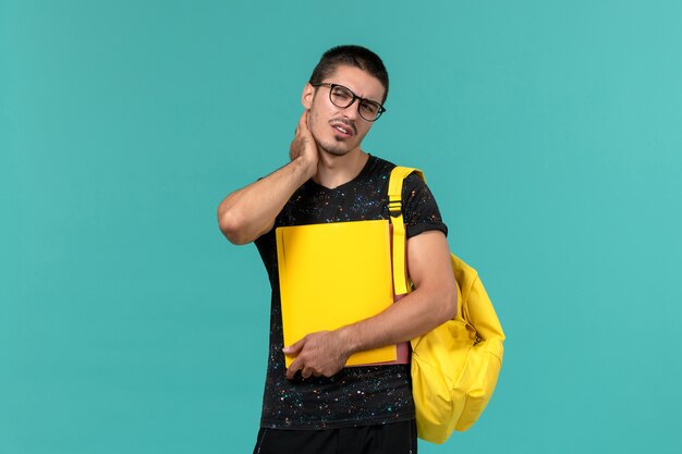 Vista frontal de um aluno do sexo masculino em uma mochila de camiseta amarela escura segurando diferentes arquivos na parede azul clara