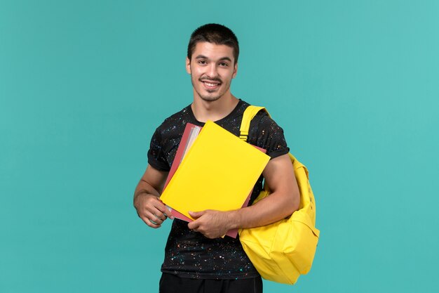 Vista frontal de um aluno do sexo masculino em uma mochila de camiseta amarela escura segurando diferentes arquivos na parede azul clara
