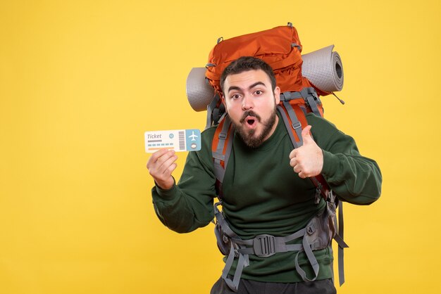Vista frontal de um cara de viagem surpreso emocional com mochila e segurando o bilhete, fazendo um gesto de ok sobre fundo amarelo