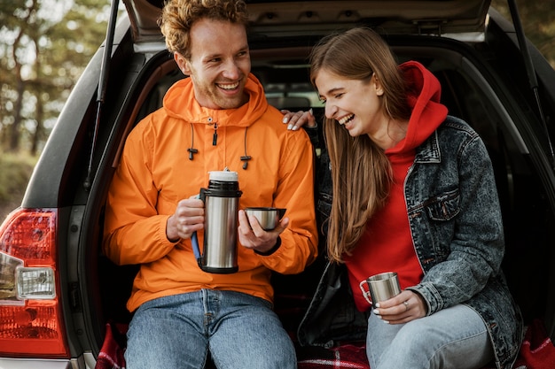 Foto grátis vista frontal de um casal sorridente desfrutando de uma bebida quente no porta-malas do carro