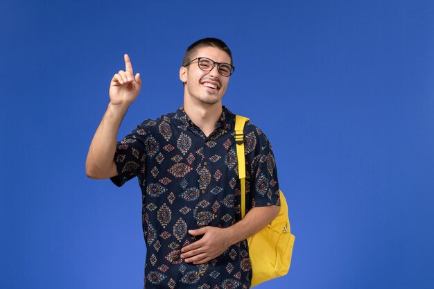 Vista frontal de um estudante do sexo masculino com camisa de algodão escura e mochila amarela posando e rindo na parede azul-clara