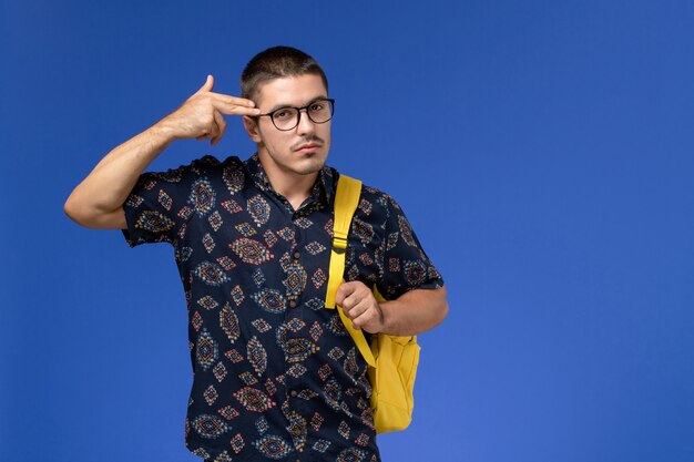 Vista frontal de um estudante do sexo masculino com camisa de algodão escura e mochila amarela, posando na parede azul claro