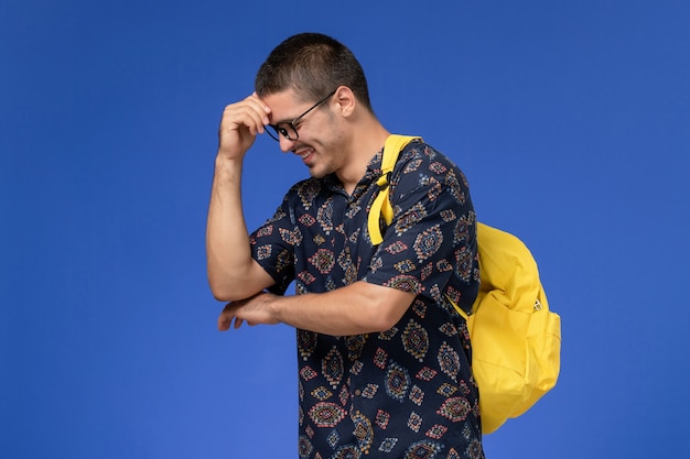 Vista frontal de um estudante do sexo masculino com camisa de algodão escura e mochila amarela rindo na parede azul clara