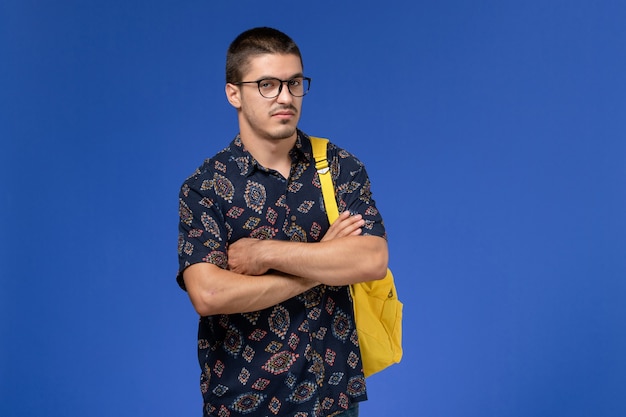 Foto grátis vista frontal de um estudante do sexo masculino com camisa de algodão escura, mochila amarela e óculos de sol ópticos, posando na parede azul