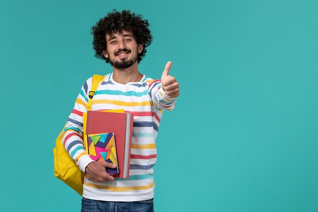 Vista frontal de um estudante do sexo masculino com camisa listrada colorida usando uma mochila amarela segurando arquivos e cadernos sorrindo na parede azul