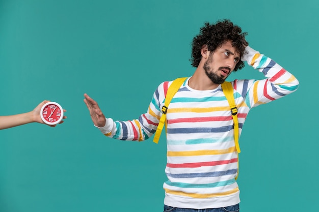 Vista frontal de um estudante do sexo masculino com camisa listrada e mochila amarela, posando na parede azul claro