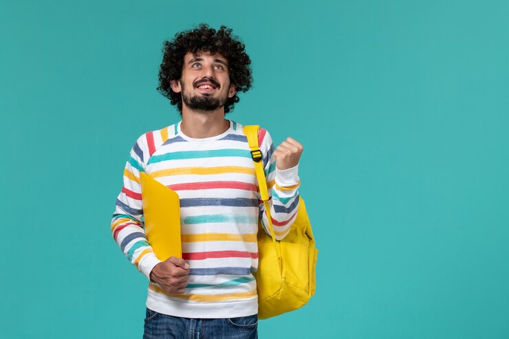 Foto grátis vista frontal de um estudante do sexo masculino com camisa listrada e mochila amarela segurando arquivos e regozijando-se na parede azul