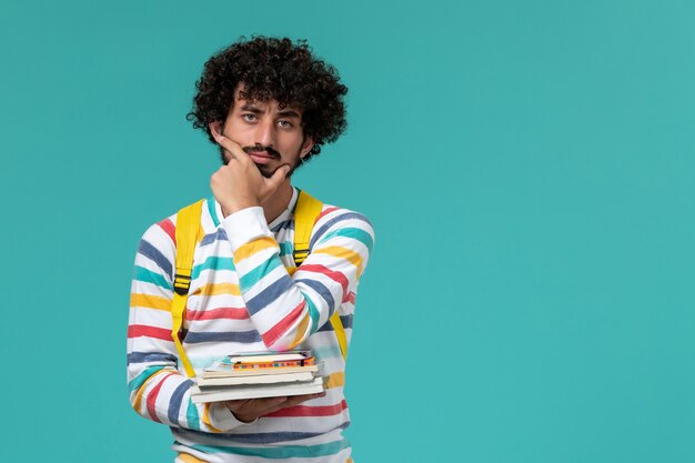 Vista frontal de um estudante do sexo masculino com camisa listrada e mochila amarela segurando livros e pensando na parede azul