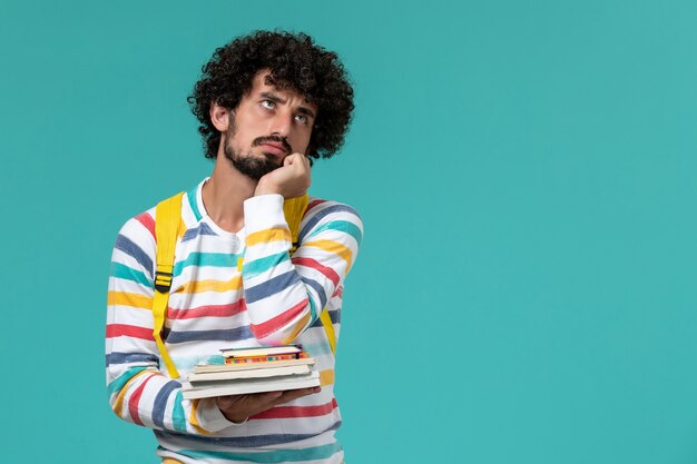 Vista frontal de um estudante do sexo masculino com camisa listrada e mochila amarela segurando livros e pensando na parede azul