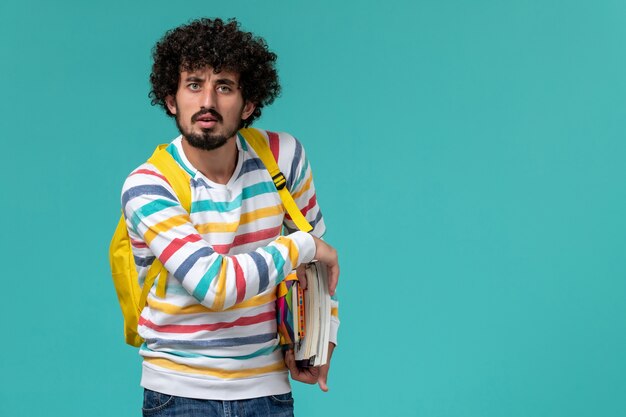 Vista frontal de um estudante do sexo masculino com camisa listrada e mochila amarela segurando livros na parede azul clara