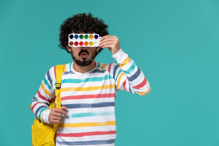 Foto grátis vista frontal de um estudante do sexo masculino com camisa listrada e mochila amarela segurando pinturas na parede azul