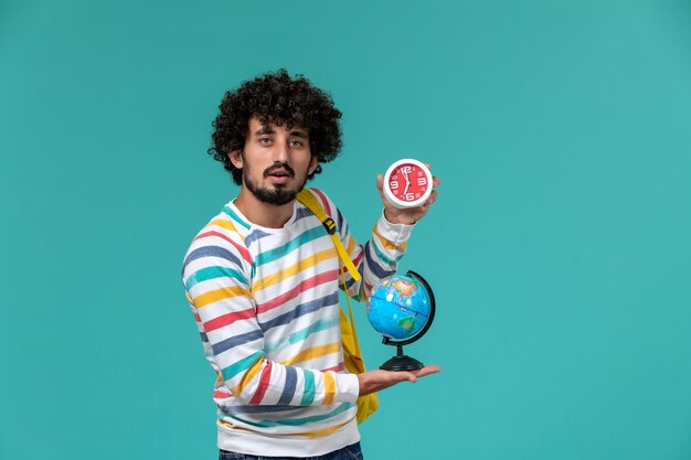 Vista frontal de um estudante do sexo masculino com camisa listrada e mochila amarela segurando um globo redondo e relógios na parede azul