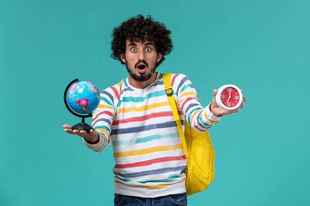 Vista frontal de um estudante do sexo masculino com camisa listrada e mochila amarela segurando um globo redondo e relógios na parede azul