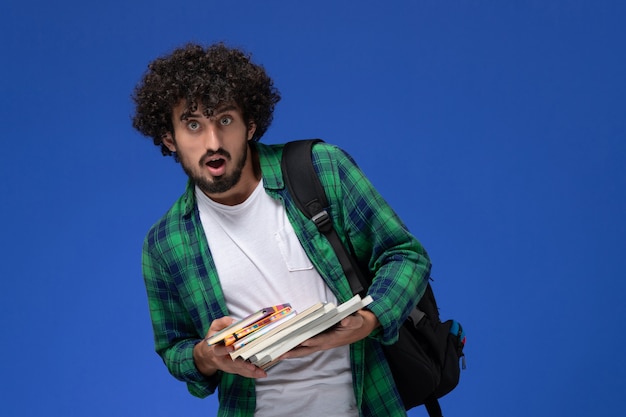 Foto grátis vista frontal de um estudante do sexo masculino com camisa quadriculada verde com mochila preta segurando cadernos e arquivos na parede azul