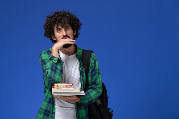 Vista frontal de um estudante do sexo masculino com camisa quadriculada verde com mochila preta segurando cadernos e arquivos na parede azul