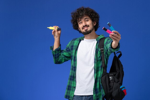 Vista frontal de um estudante do sexo masculino com camisa quadriculada verde com mochila preta segurando canetas de feltro sorrindo na parede azul